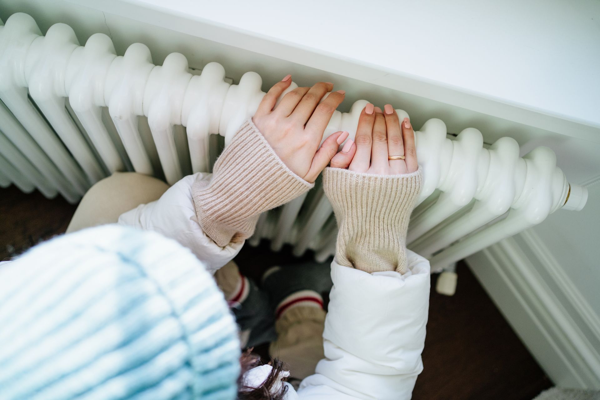 Person wearing a hat and gloves, warming hands on a white radiator.