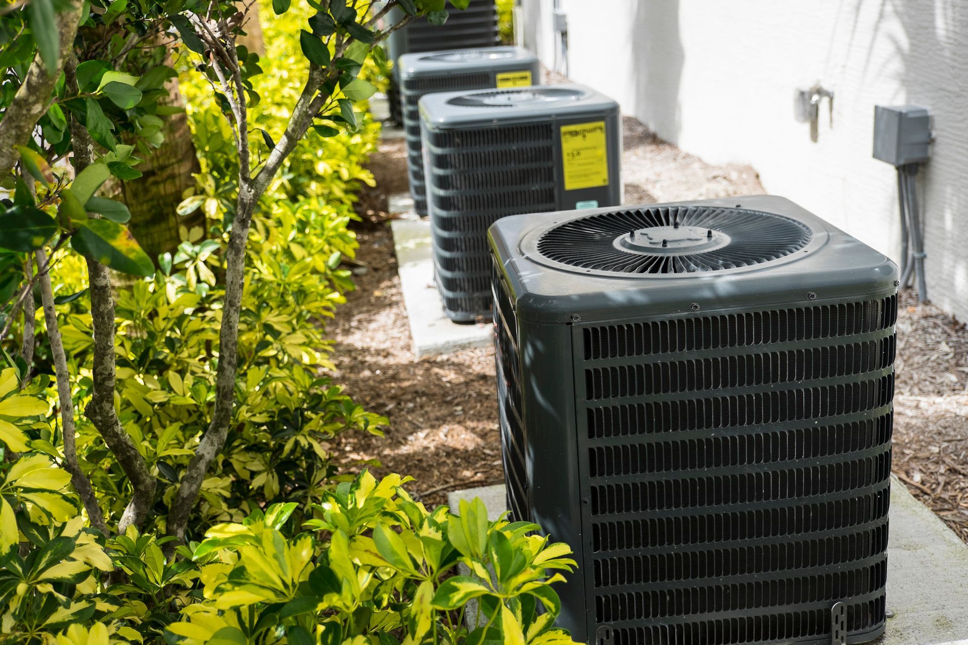 Row of dark gray air conditioning units next to green bushes and a white building.