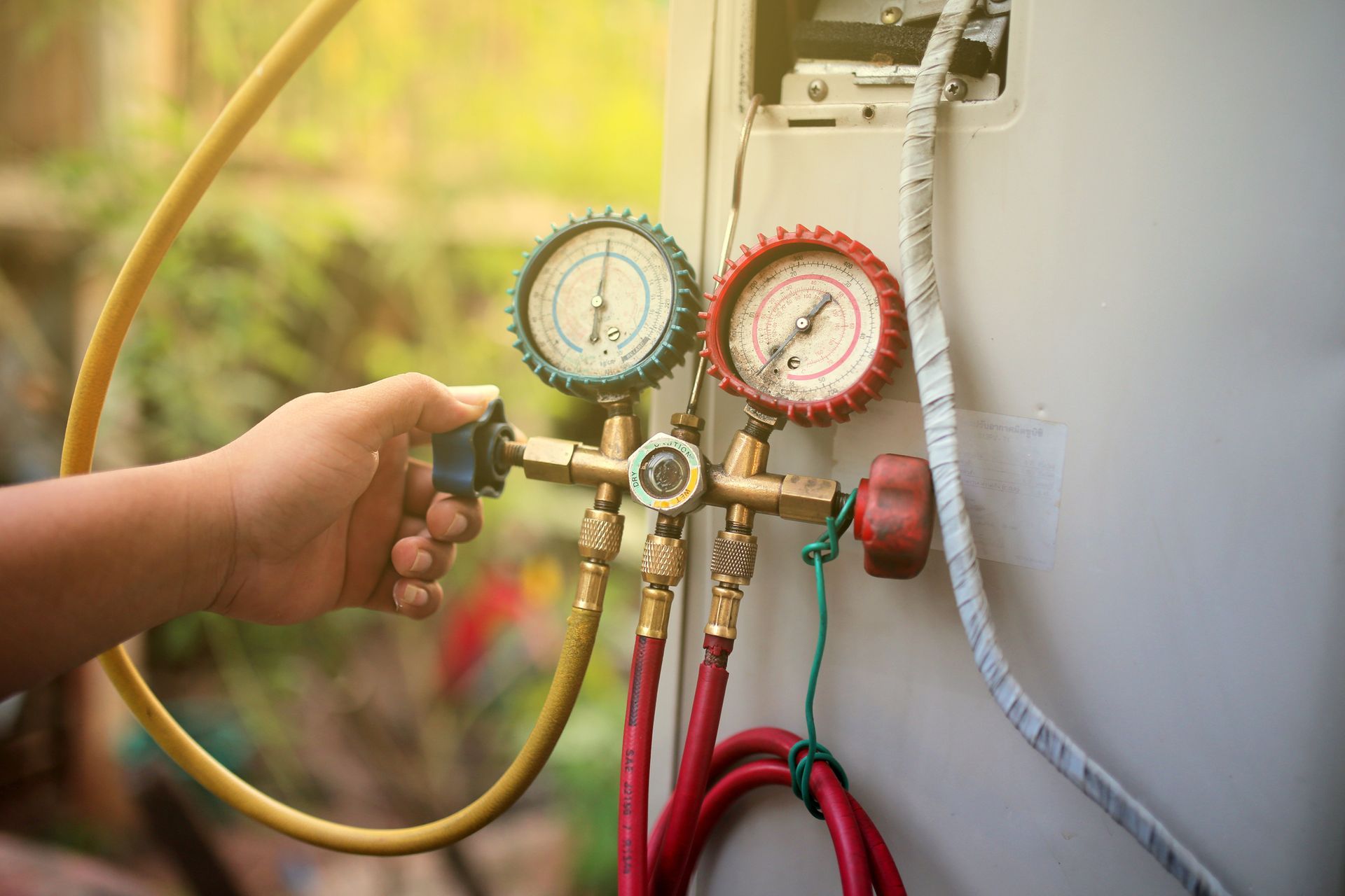 Hand adjusting gauges on an AC unit outdoors, connected by yellow and red hoses.