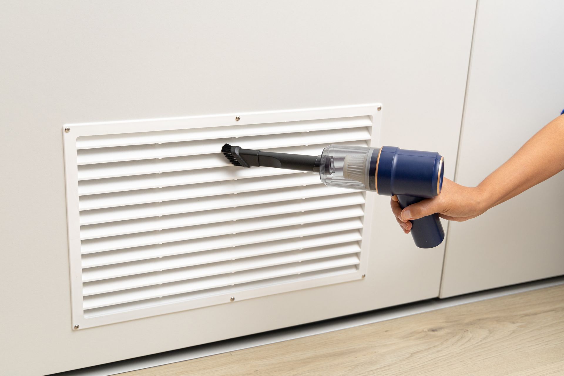 Hand using a blue handheld vacuum to clean a white air vent.