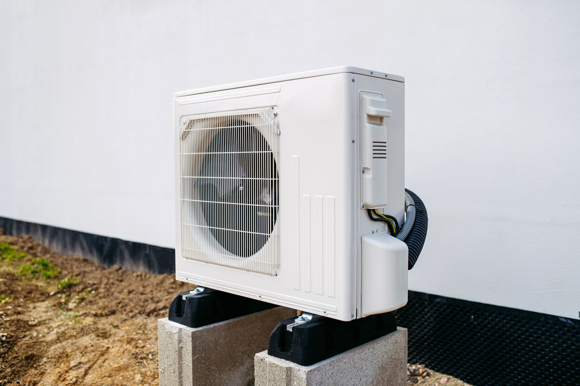 Air conditioner unit, white, mounted on concrete blocks outside against a white wall.