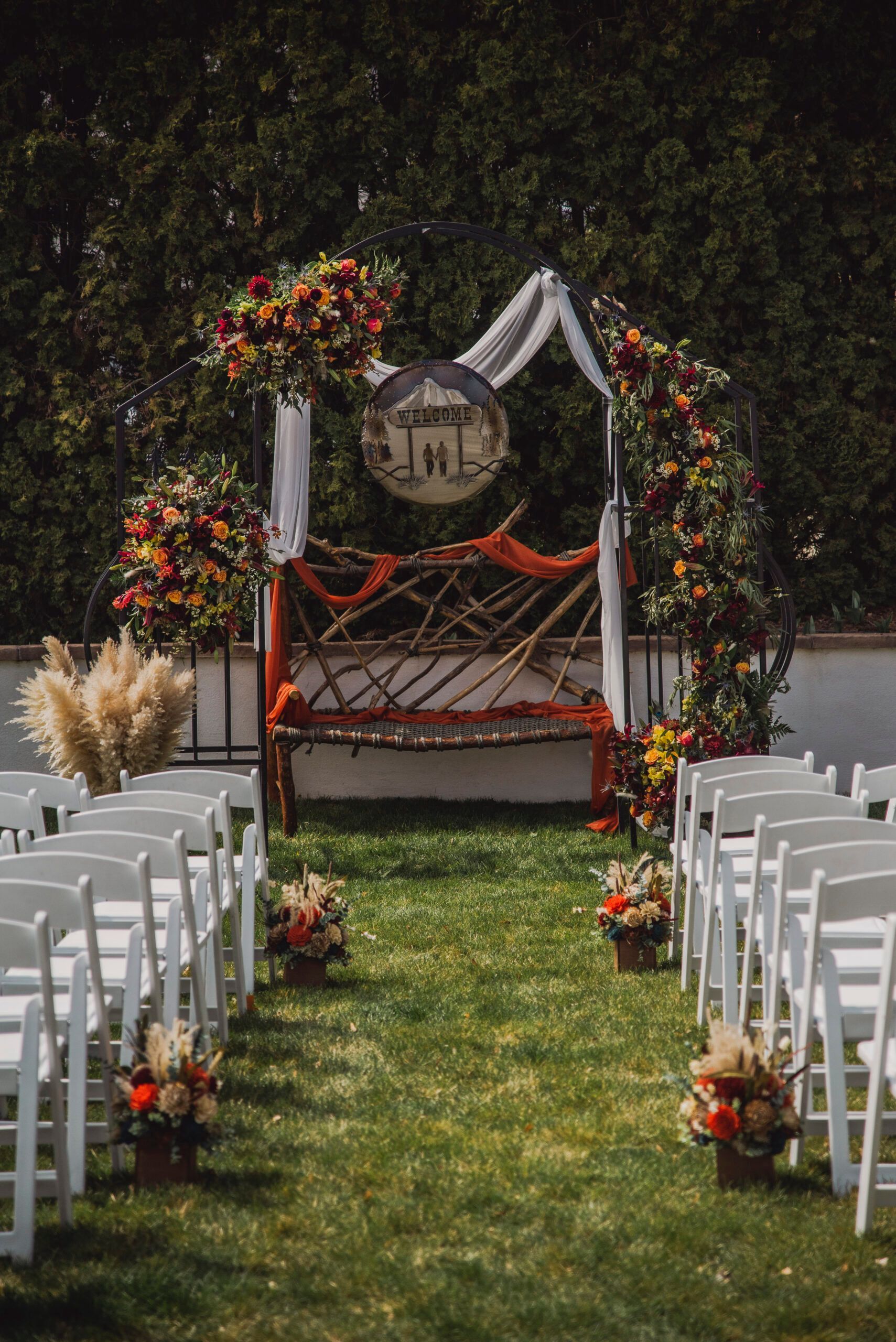 A row of white chairs are lined up in front of a wedding arch decorated with flowers.