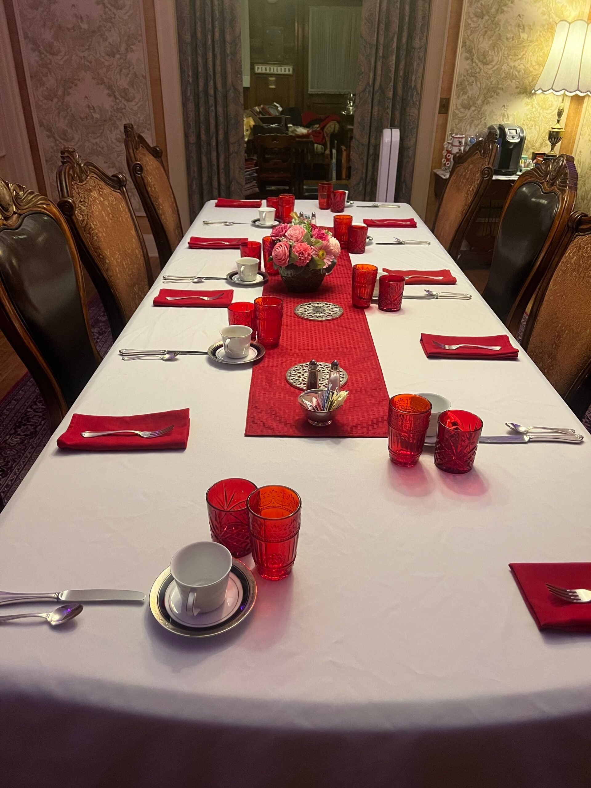 A long table with red glasses , cups , and silverware on it.
