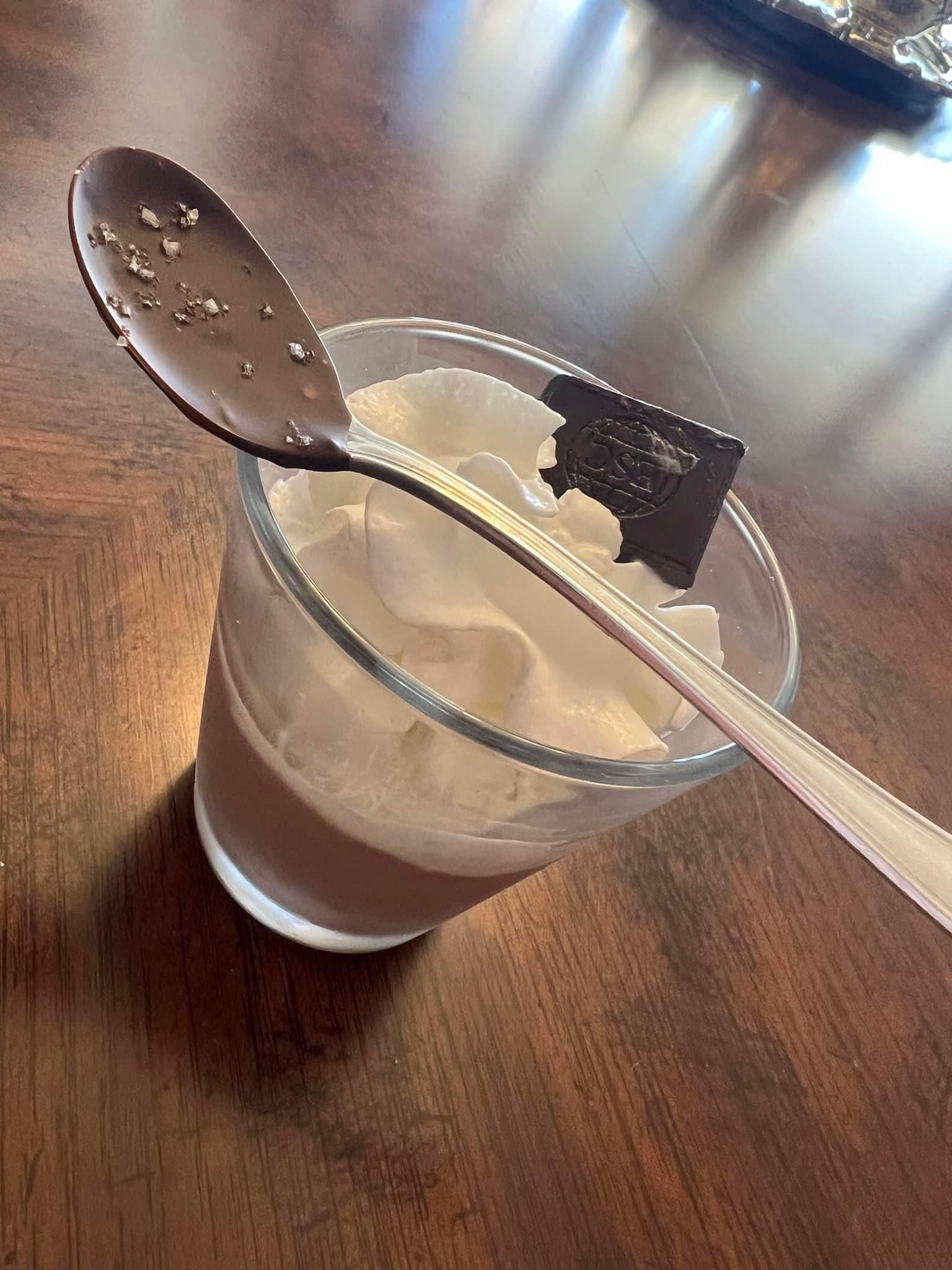 A glass of chocolate pudding with whipped cream and a chocolate spoon on a wooden table.