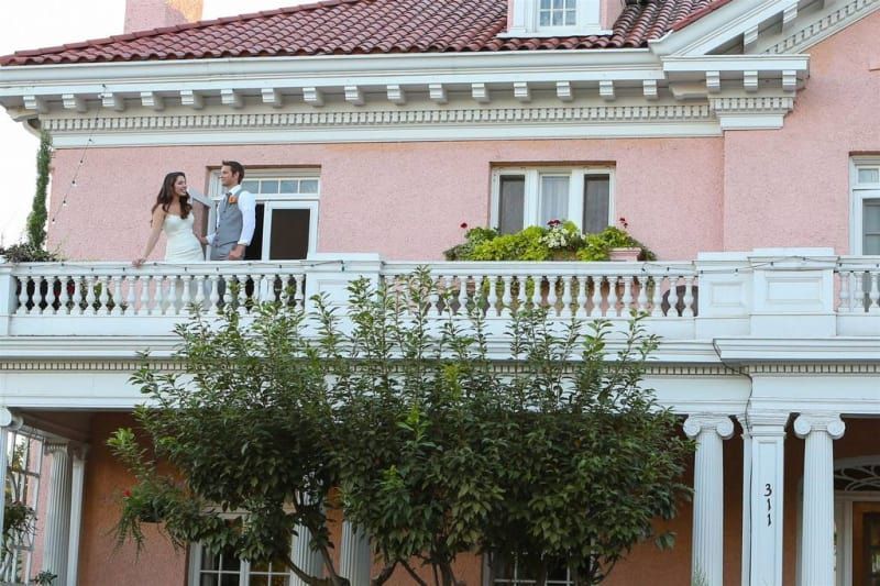 A bride and groom are standing on a balcony of a large pink house.