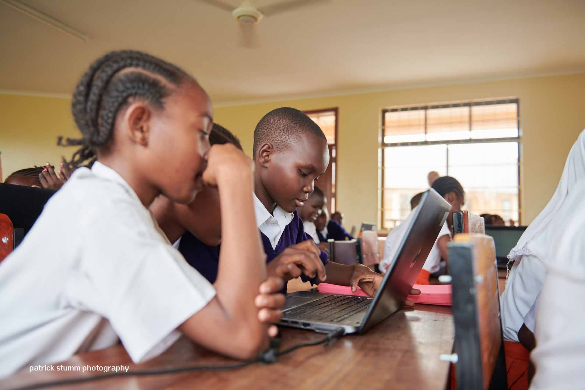Eine Gruppe Kinder in orangefarbenen Schuluniformen, einige winken. Drinnen, wahrscheinlich in einer Schule.