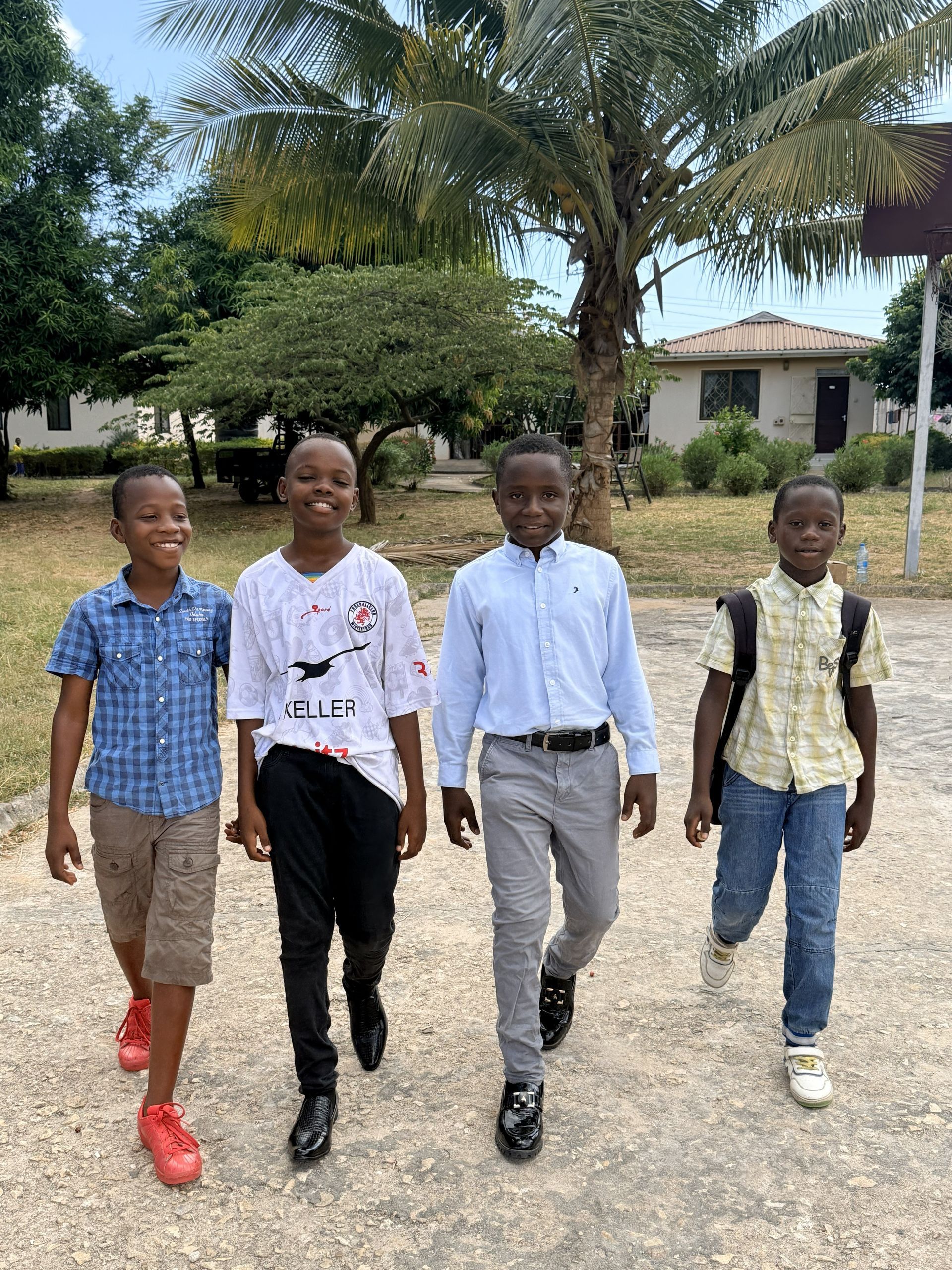 Vier junge Leute gehen draußen auf die Kamera zu; im Hintergrund sind Gebäude und Bäume zu sehen.