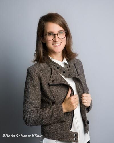 Junge Person mit geflochtenen Haaren lächelt, im Freien, weicher Hintergrund.