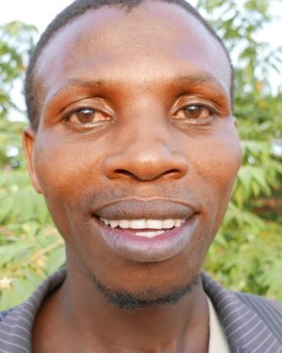 Eine Frau mit dunklen Haaren lächelt und trägt ein rosa Hemd mit Kragen. Im verschwommenen Hintergrund ist grünes Laubwerk zu sehen.