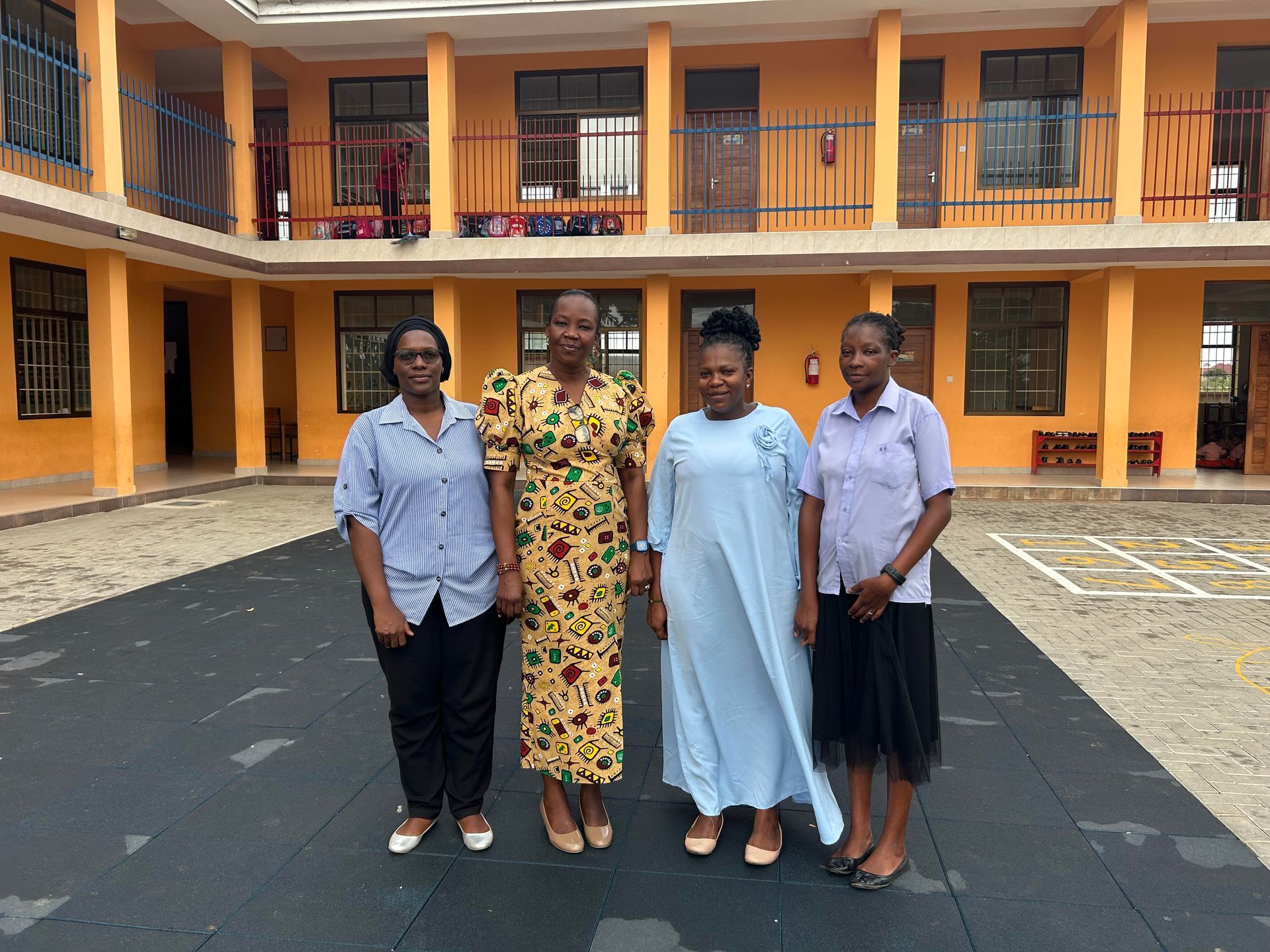 Ein Schulhof mit mehreren gelben Bussen, die vor orangefarbenen, zweistöckigen Schulgebäuden unter bewölktem Himmel parken.