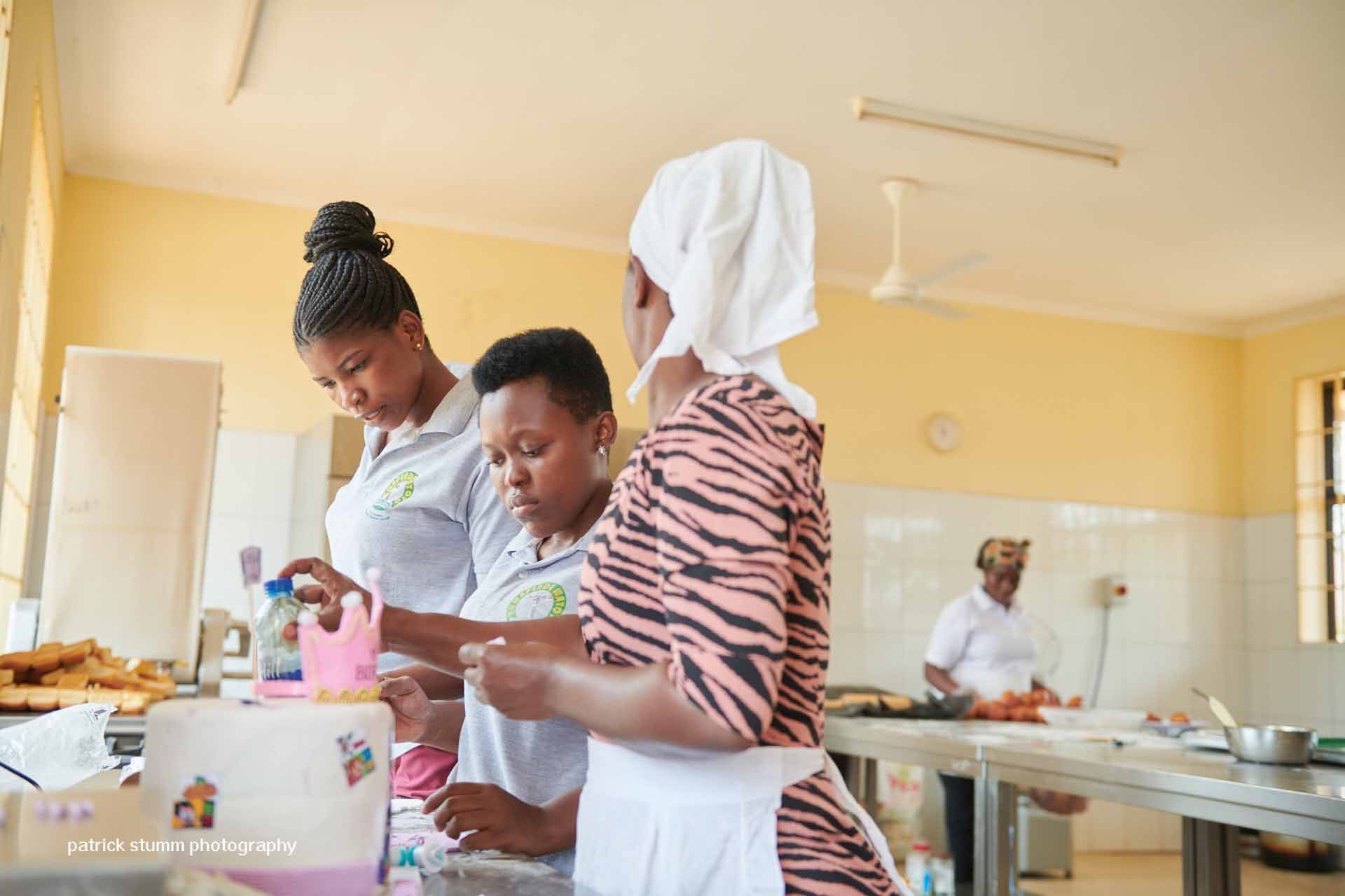 Drei Frauen bereiten in einer Küche Essen zu. Eine trägt ein Kopftuch, die anderen tragen Schürzen. Hell erleuchteter Raum.