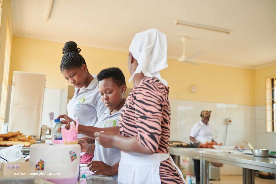 Drei Frauen bereiten in einer Küche Essen zu. Eine trägt ein Kopftuch, die anderen tragen Schürzen. Hell erleuchteter Raum.