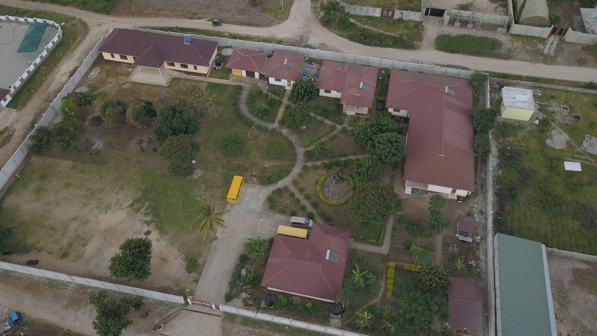 Luftaufnahme einer Schule mit mehreren Gebäuden und einem gelben Schulbus, umgeben von einem Zaun und einem Feldweg.