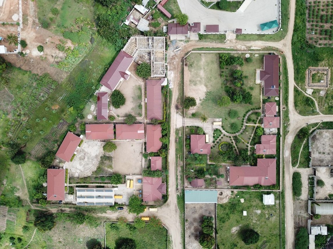 Luftaufnahme eines Komplexes mit rotgedeckten Gebäuden, einer zentralen Straße und grüner Vegetation in ländlicher Umgebung.