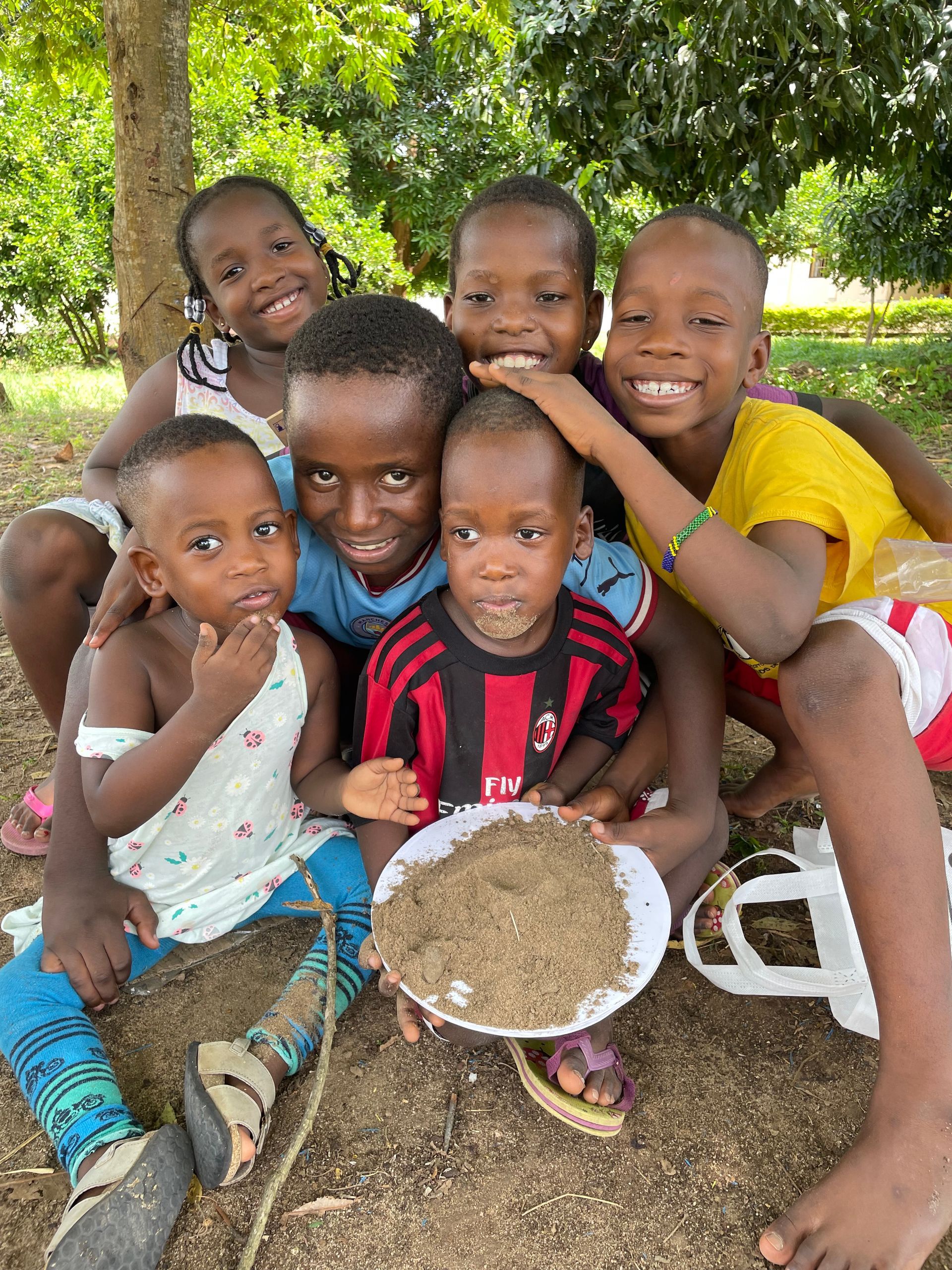 Eine Gruppe lächelnder Kinder im Freien, einige halten einen Teller mit Essen in den Händen und sitzen unter einem Baum auf dem Boden.