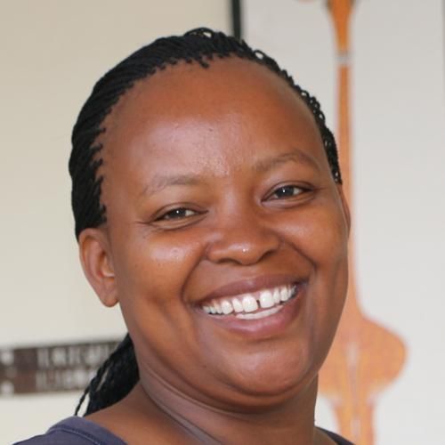 Eine Frau mit dunklen Haaren lächelt und trägt ein rosa Hemd mit Kragen. Im verschwommenen Hintergrund ist grünes Laubwerk zu sehen.