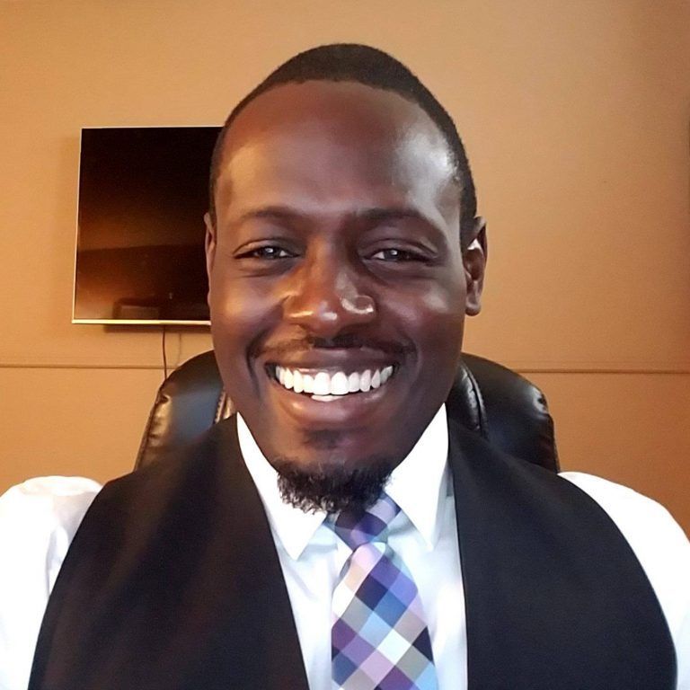 Man in black vest, white shirt, and patterned tie smiles, seated in front of a tv screen.