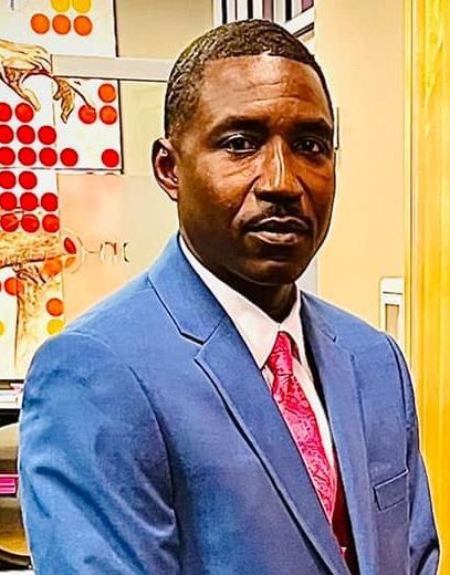Man in blue suit and pink tie; posed in an office setting, looking at the camera.
