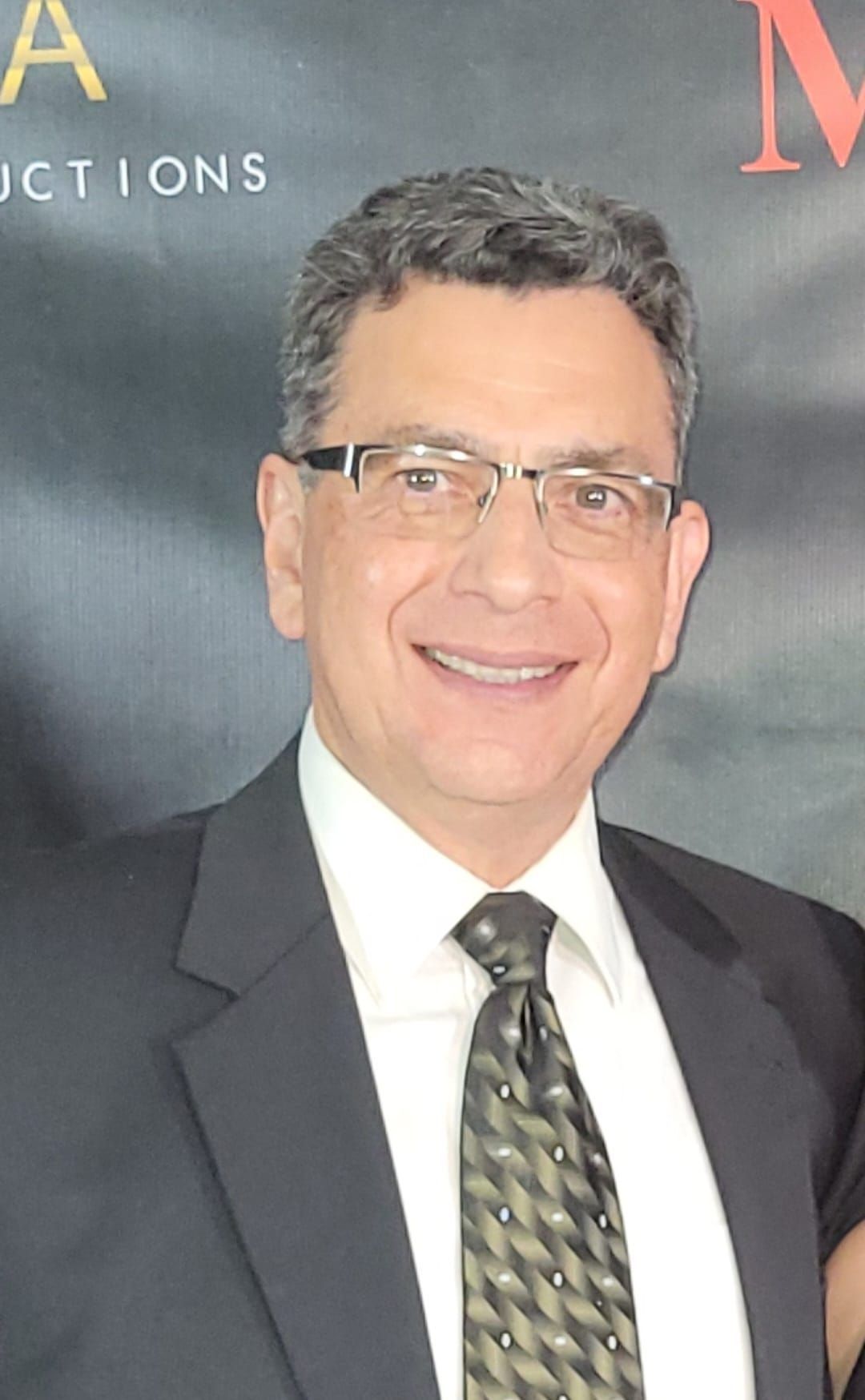 Man in a suit smiles, wearing glasses, in front of a dark backdrop.