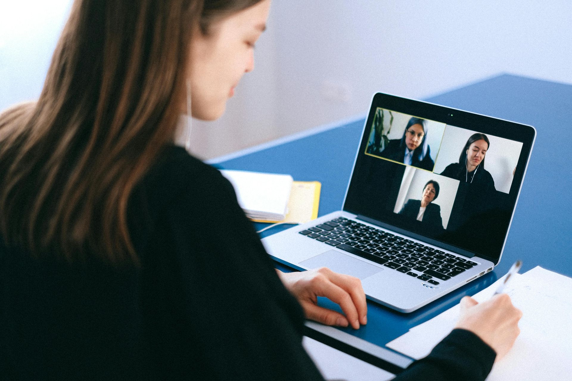 Woman on a video call, looking at laptop screen with three other people. She is taking notes on paper.