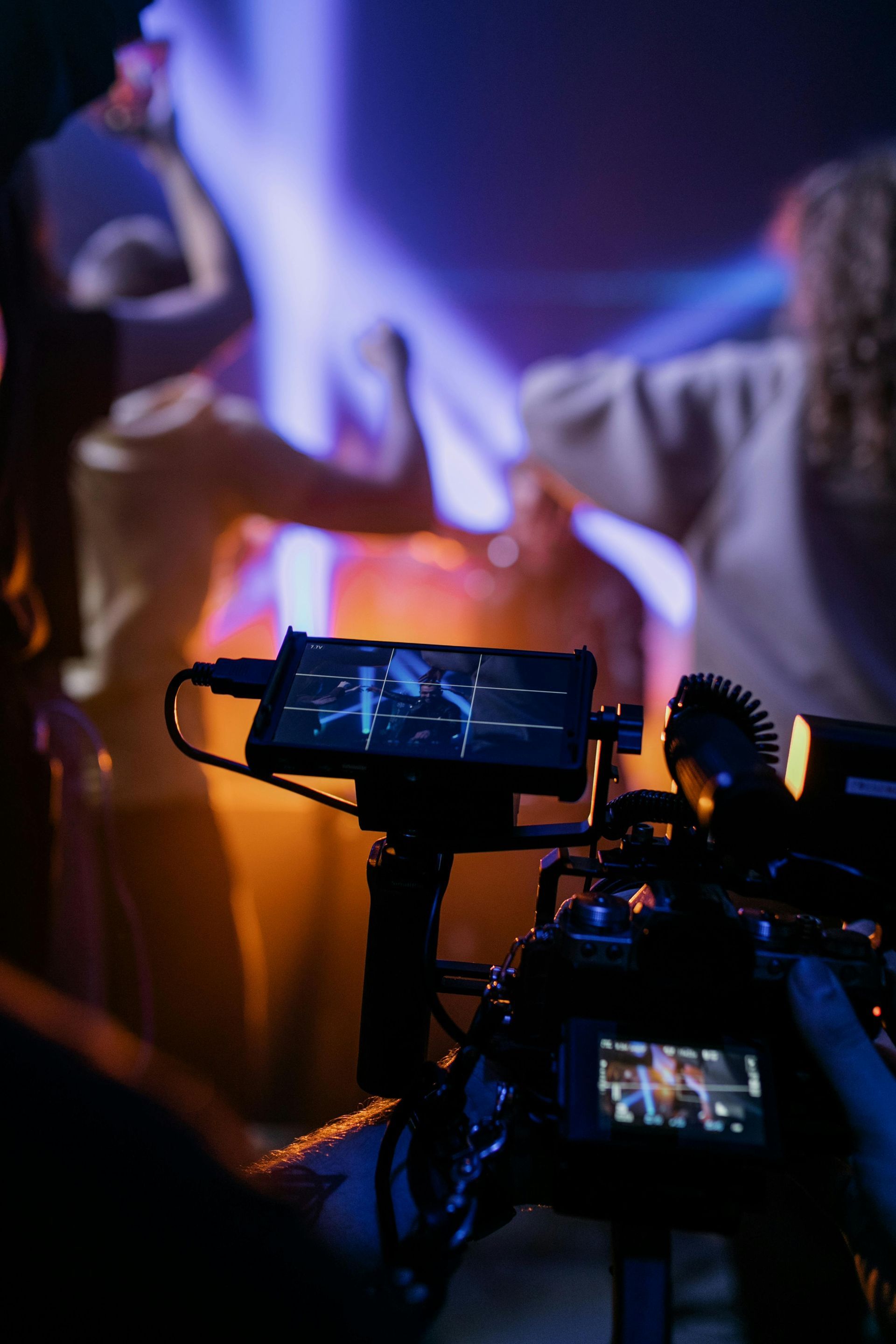 A video camera recording dancers on stage, illuminated by orange and blue stage lights.