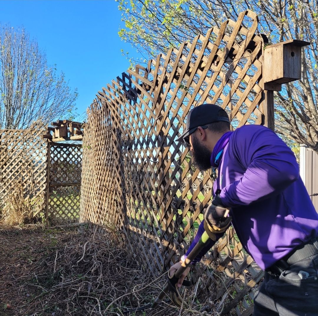 A man in a purple shirt is standing in front of a wooden fence holding a shovel.