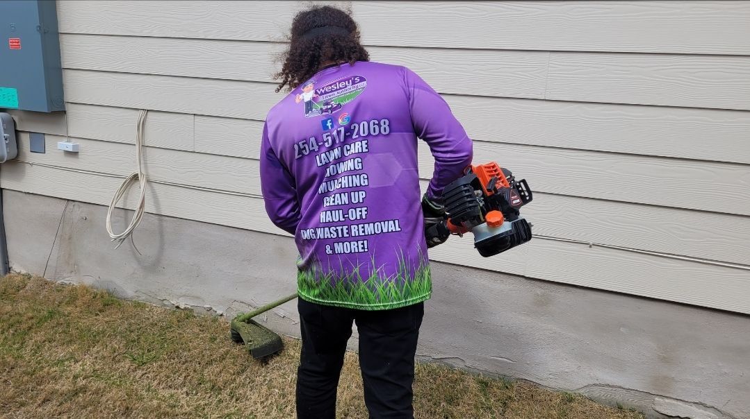 A man in a purple shirt is standing in front of a house holding a lawn mower.