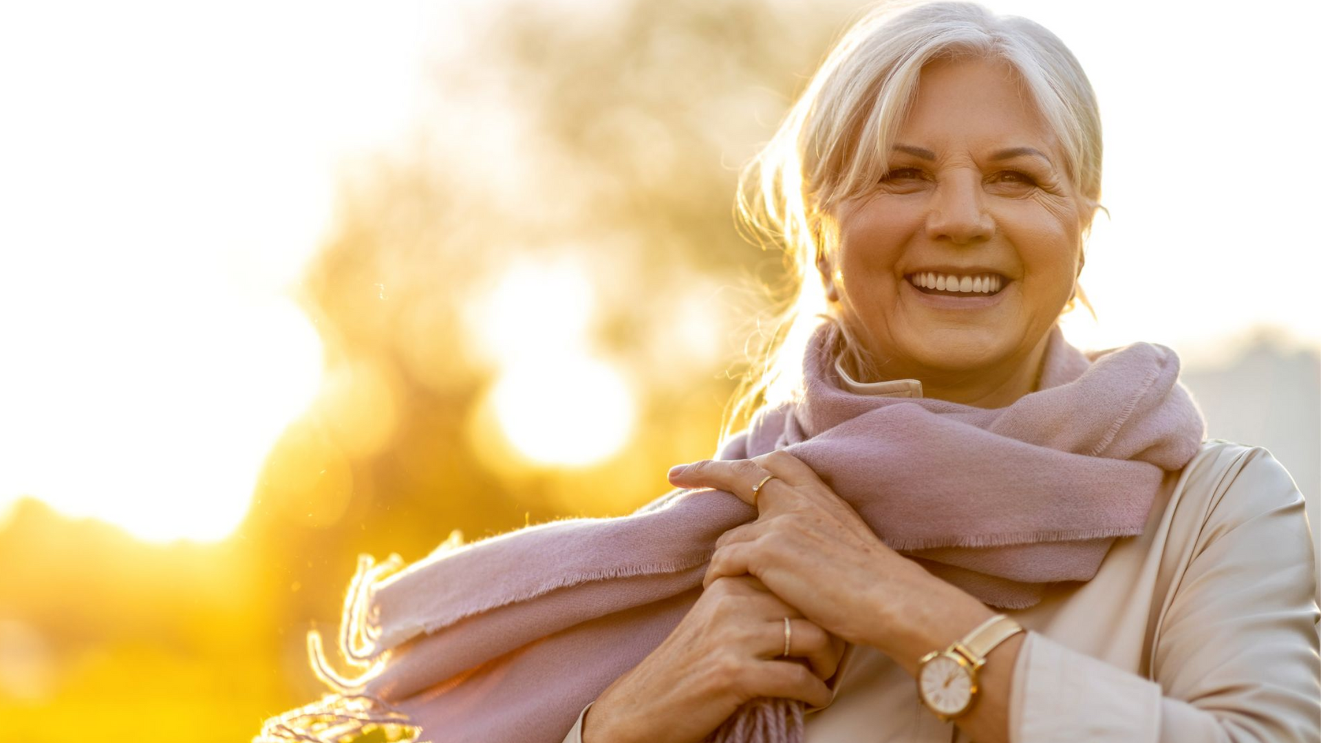 Een glimlachend persoon, gehuld in een zachte paarse sjaal, wordt verlicht door een warme, gouden zonsondergang in een buitenomgeving.
