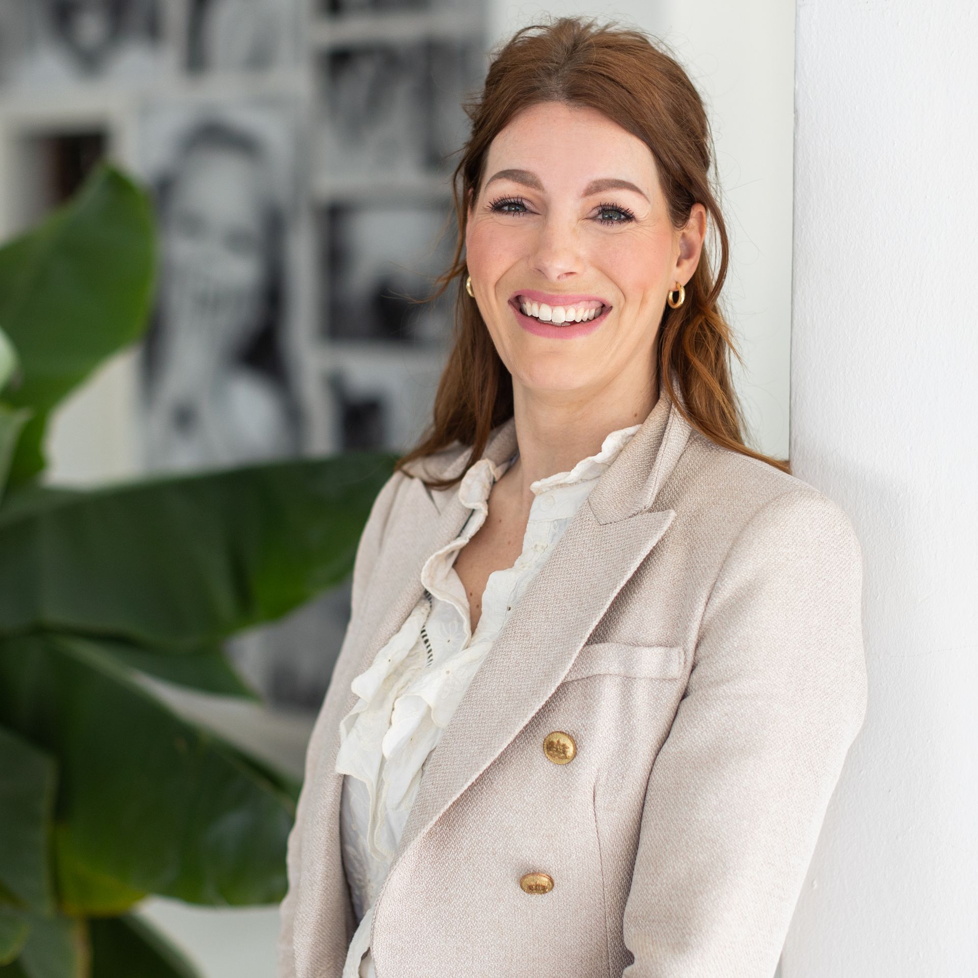 Een vrouw in een lichtgekleurde blazer glimlacht terwijl ze poseert bij een plant en een muur.