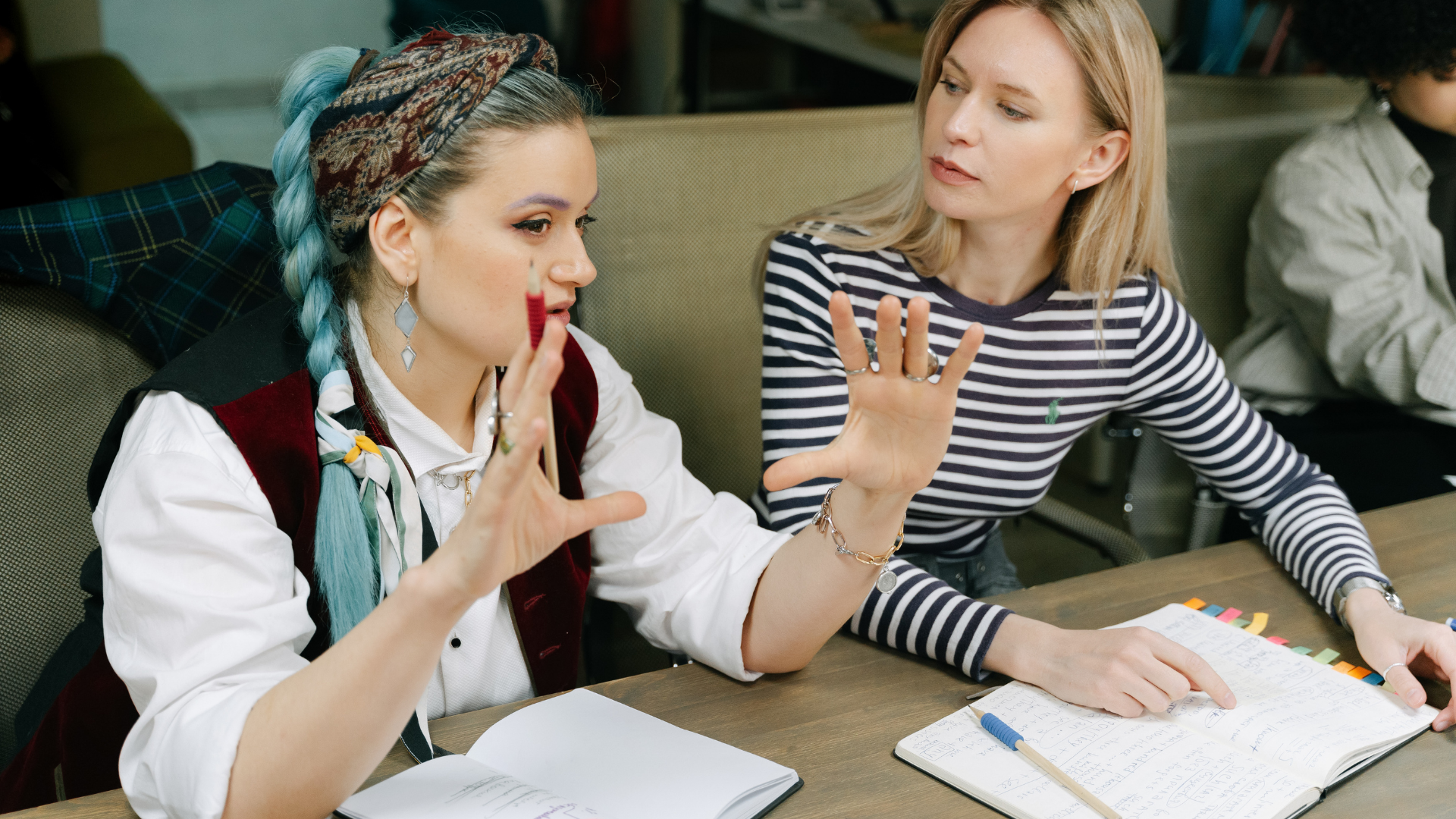 Two people sit at a table with open notebooks, discussing a project in an office setting.