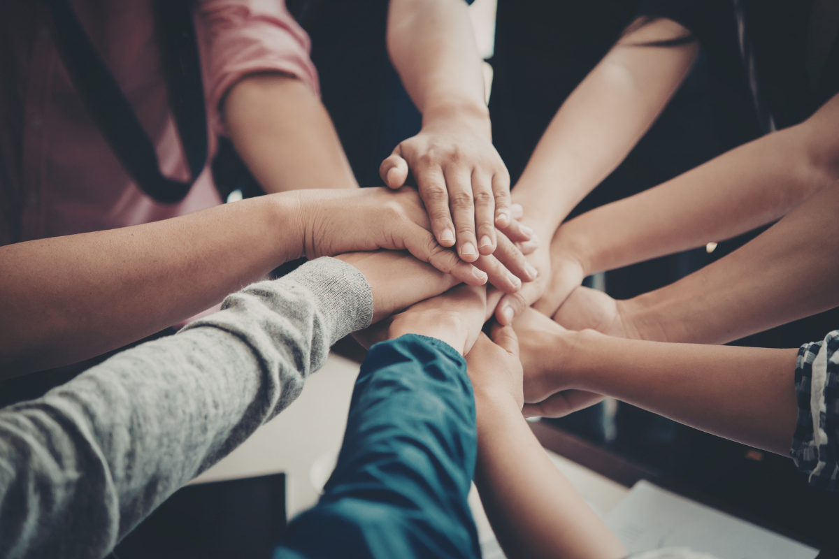 A group of people place their hands together in the center in a gesture of unity, teamwork, and collaboration.