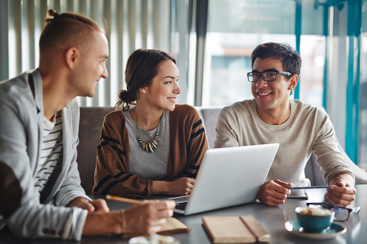 Three people sit around a laptop in a bright, modern office space, smiling while engaged in a collaborative discussion.