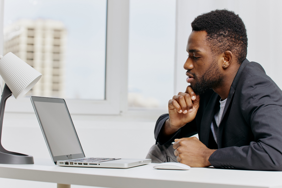 A person in a dark suit sits at a white desk, thoughtfully looking at a laptop screen in a bright office.