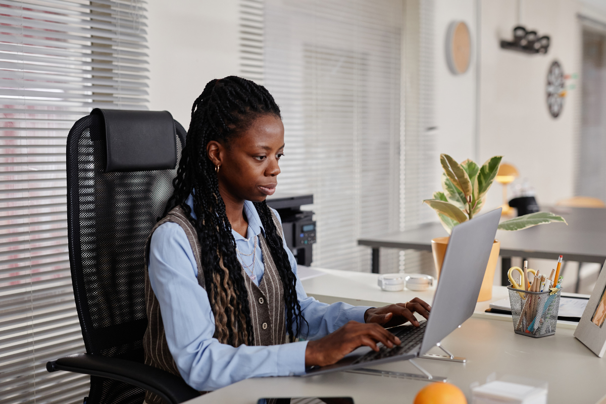 A person with long braided hair wearing a collared shirt and vest works on a laptop at a desk in an office.