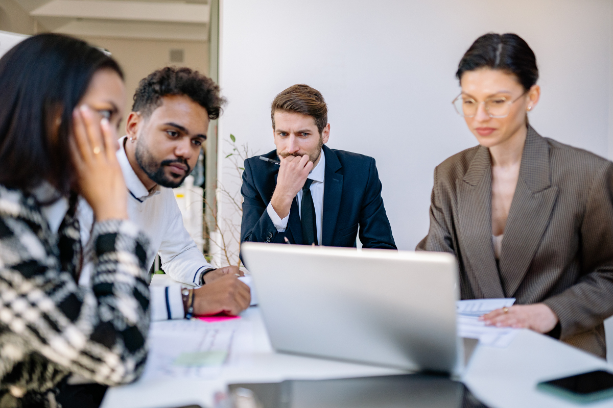 Four professionals sit around a white table looking at a laptop during a collaborative office meeting.