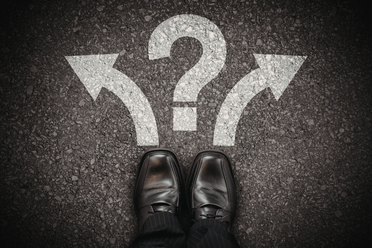 A pair of dark shoes stands on asphalt facing a painted white question mark between two arrows pointing left and right.