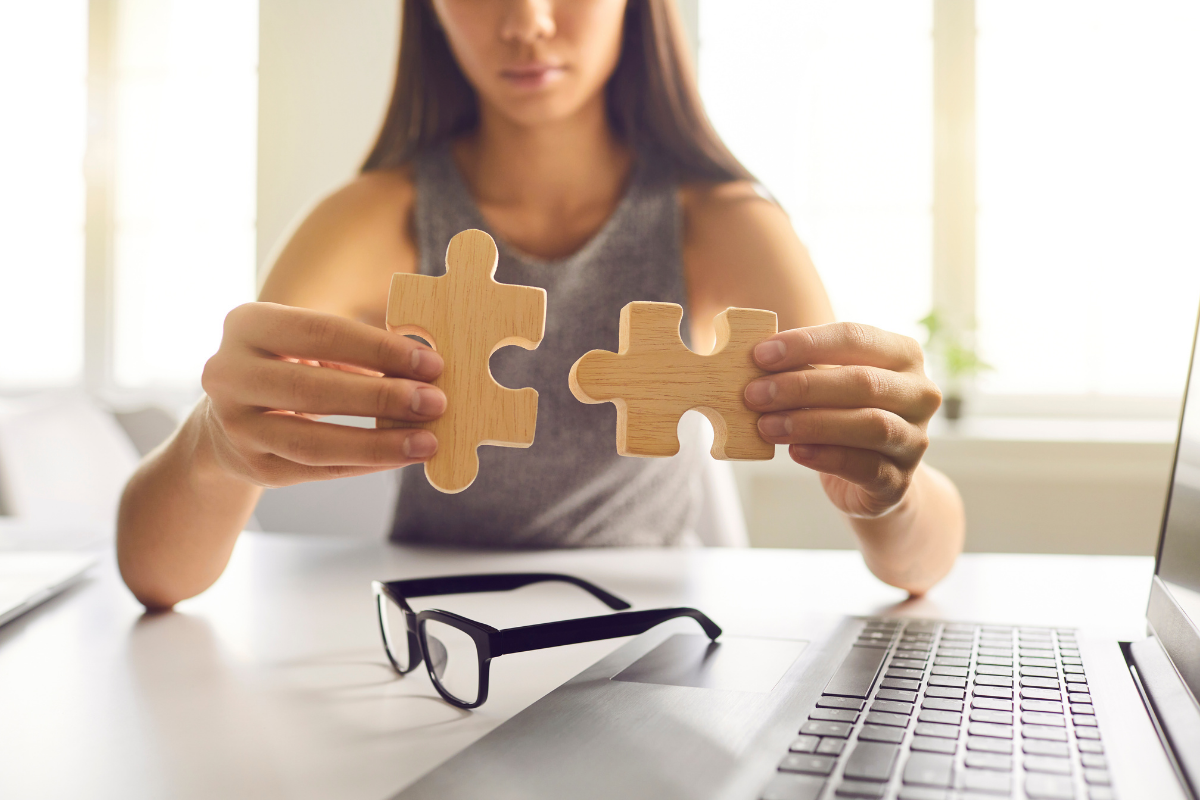 A person sitting at a desk with a laptop and glasses holds two wooden puzzle pieces together.