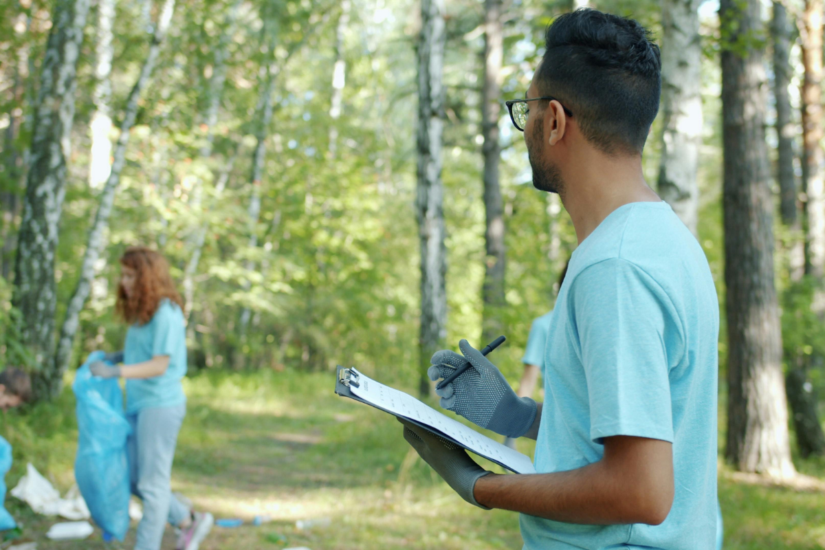 A volunteer in a blue t-shirt and gloves holds a clipboard in a forest, while another person collects trash in the back.