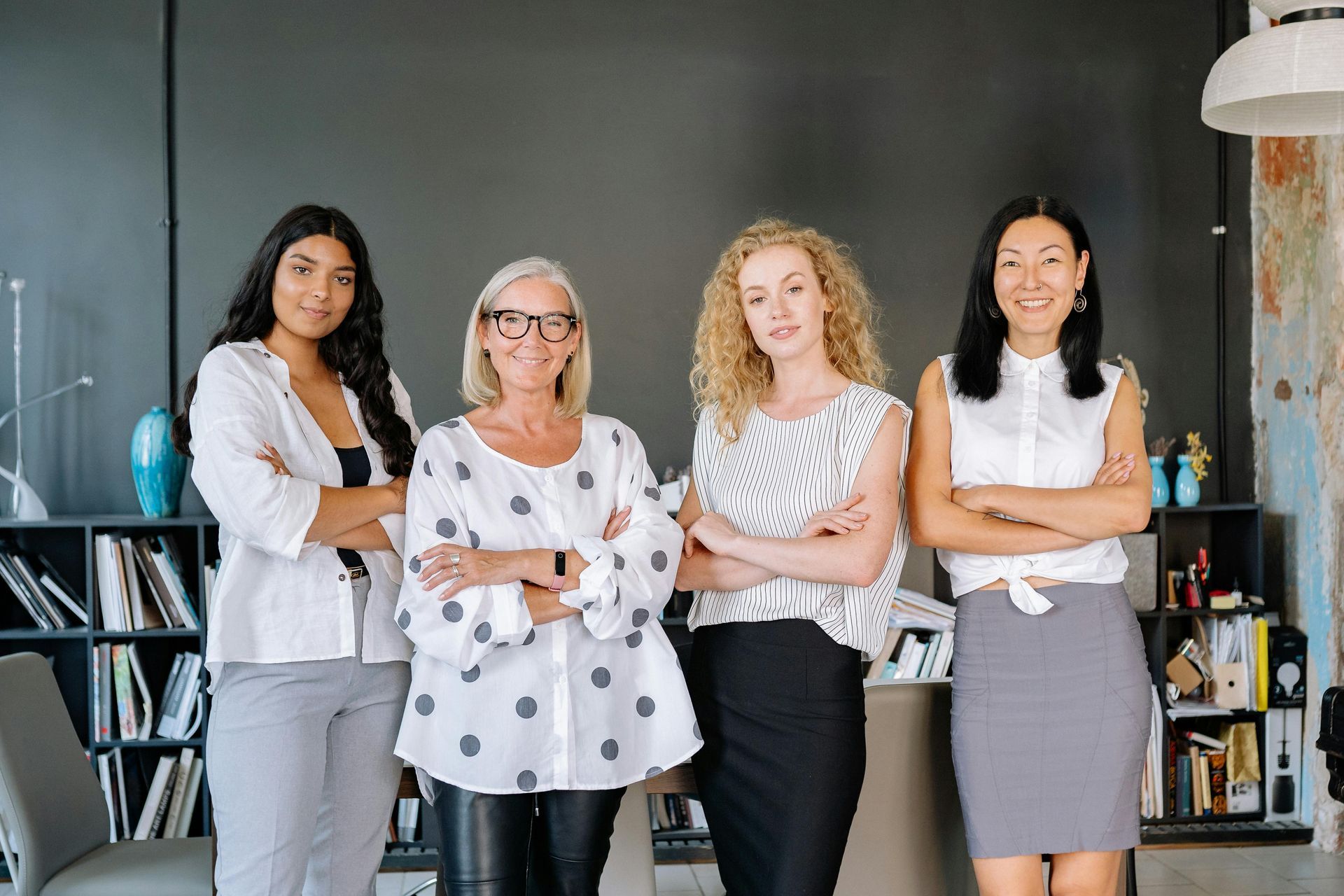 Four professionals with arms crossed standing in a modern office space with dark walls and shelving units.