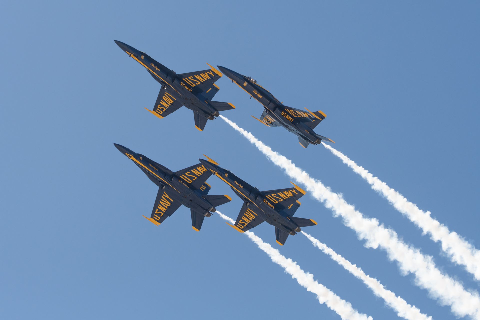 Four blue and gold US Navy Blue Angels jets flying in formation against a clear blue sky, leaving white contrails.