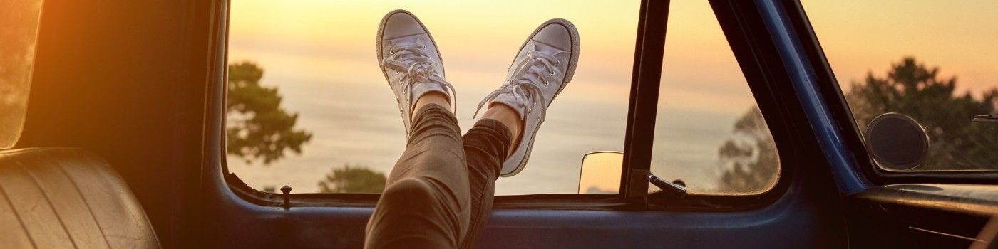 Person's legs resting on car window. Sunset over the ocean. Wearing white sneakers.