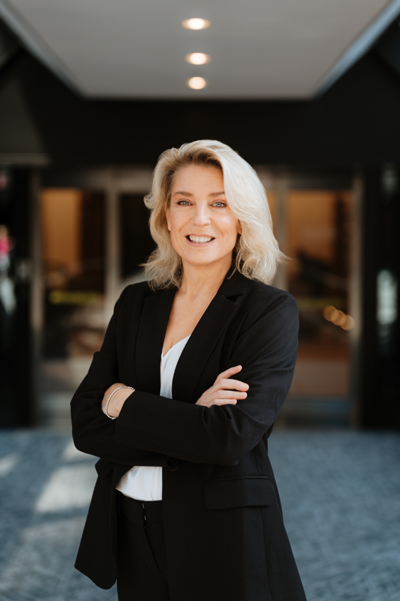 A woman in a black suit is standing with her arms crossed in front of a building.