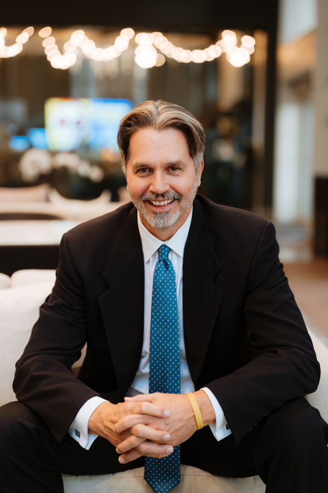 A man in a suit and tie is sitting on a couch with his hands folded.