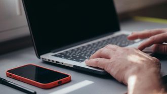 A person is typing on a laptop computer next to a cell phone.