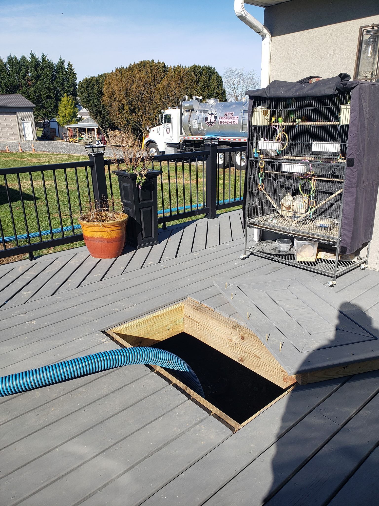 Blue hose extends from open deck access. Gray deck with black railing, potted plant, and equipment. Bright day.