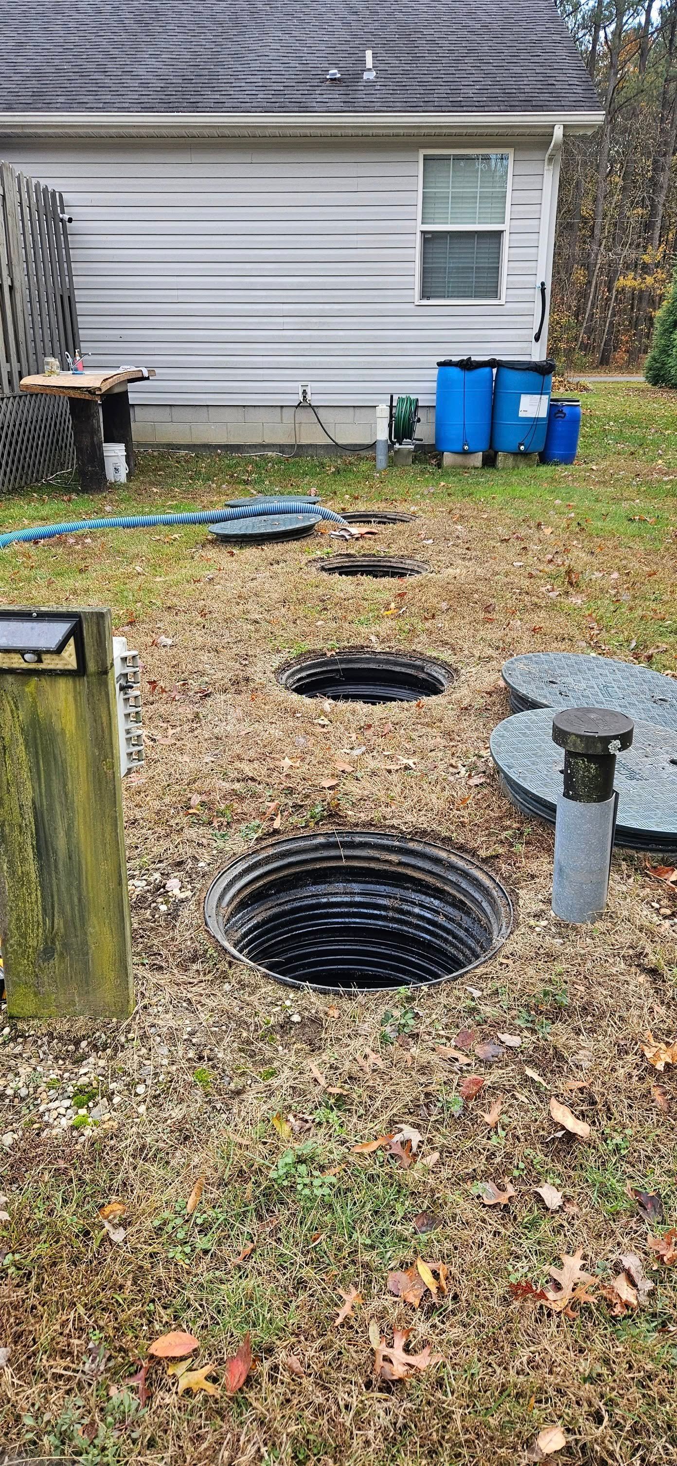 Open septic tank surrounded by gravel, in a grassy yard, near a white building.