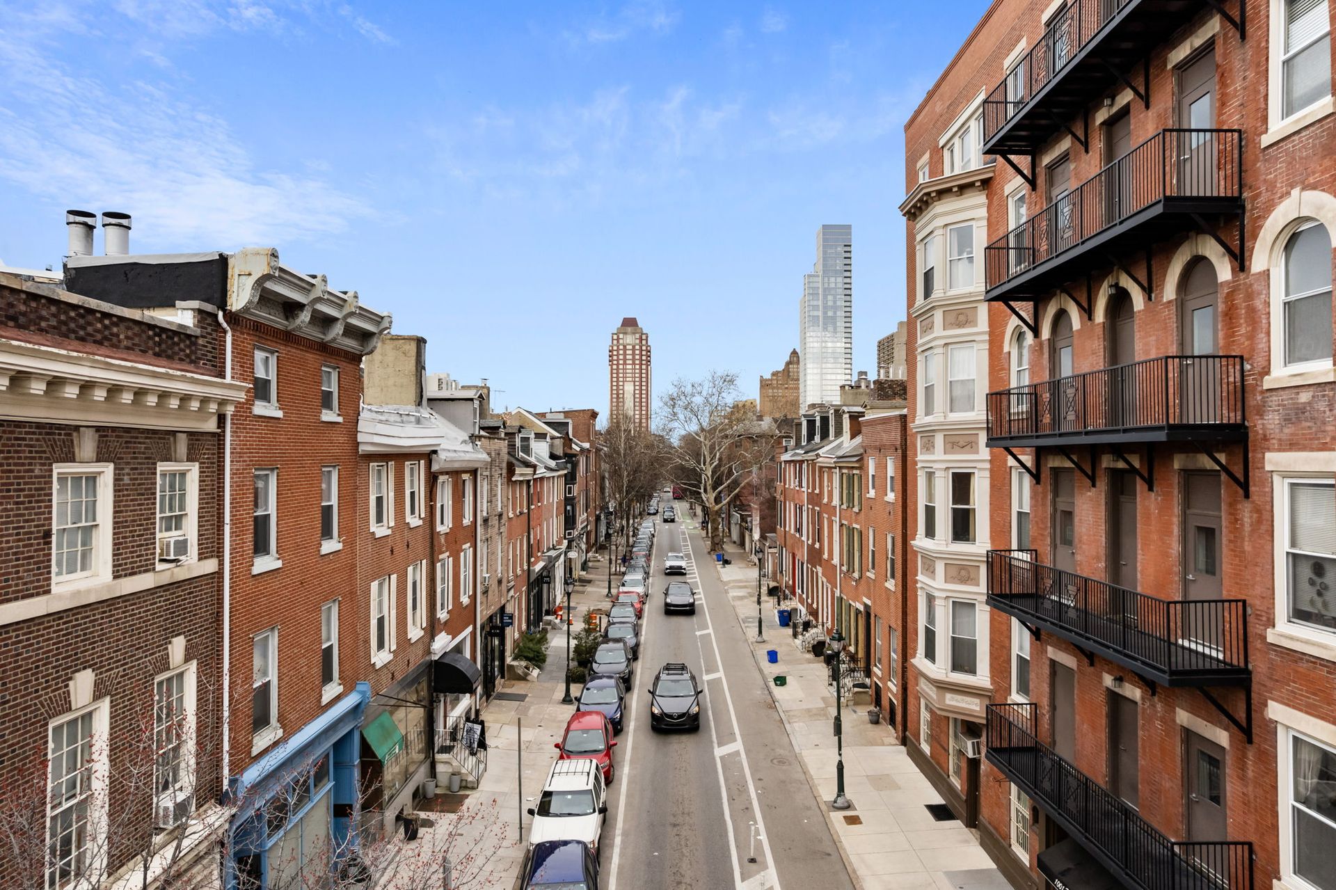 A street view of a city lined with multi-story brick residential buildings, parked cars, and a distant tower.