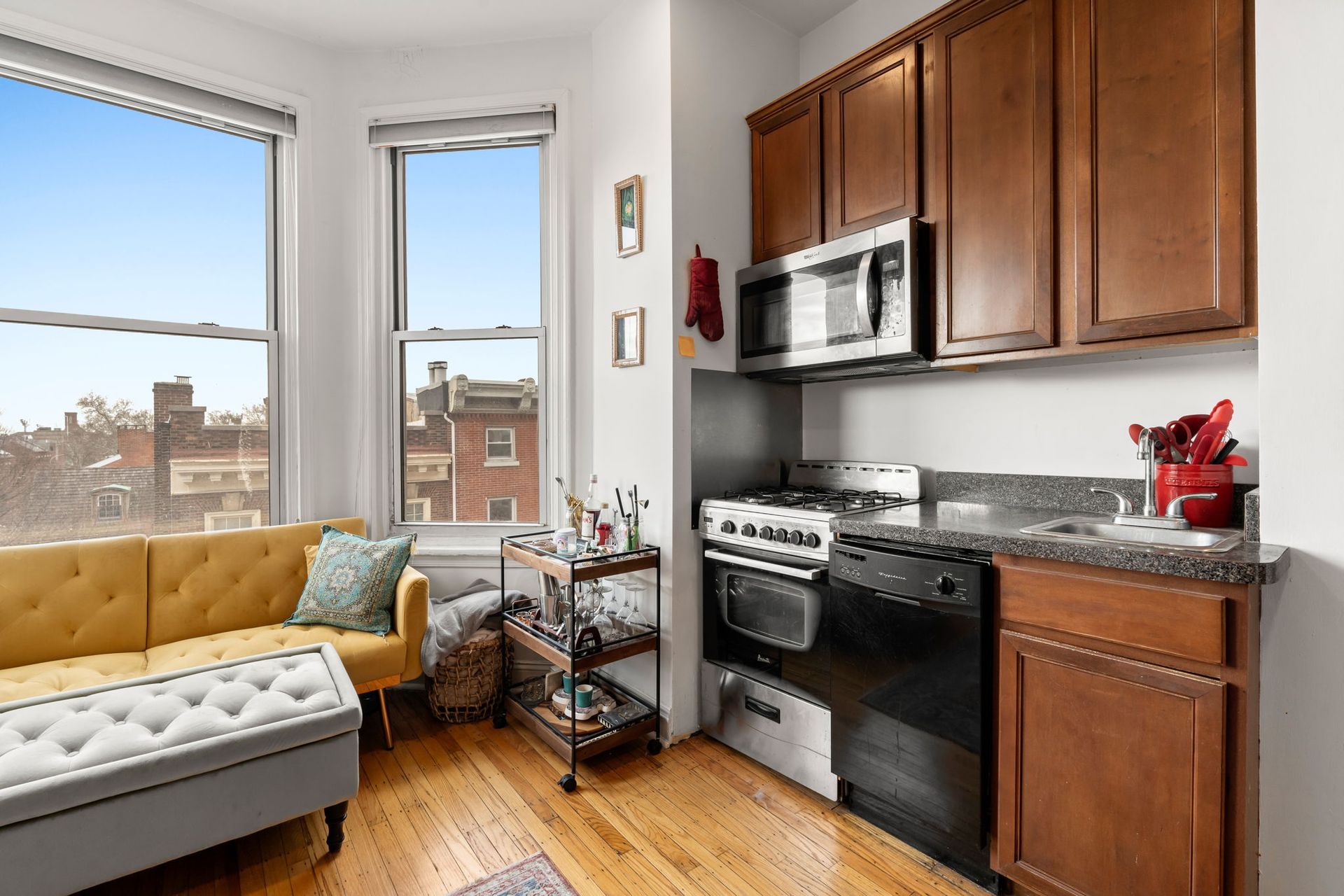 A compact apartment space featuring a yellow sofa, gray ottoman, rolling cart, and a kitchen with brown cabinets.