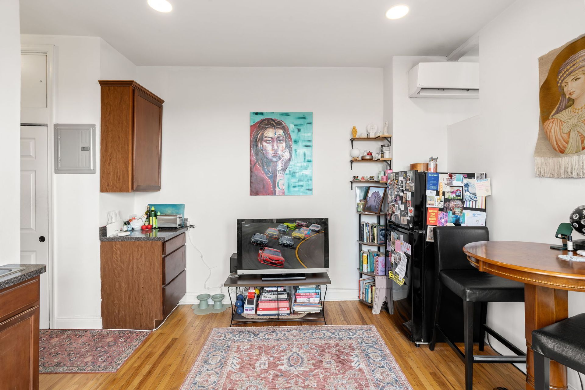 A studio apartment with a kitchen area, a television on a stand, a bookshelf next to a refrigerator, and a small table.