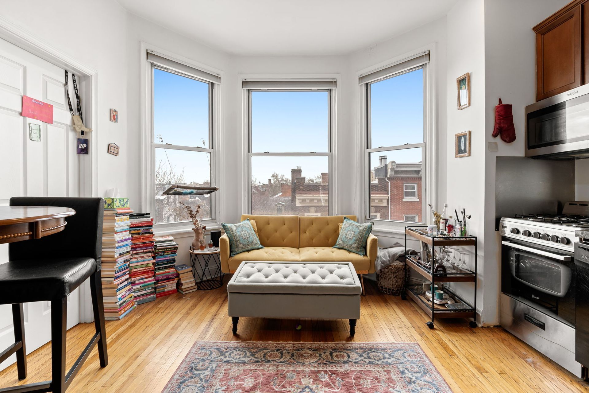 A cozy living area featuring a yellow sofa, an ottoman, and a large stack of books in a sunlit bay window room.